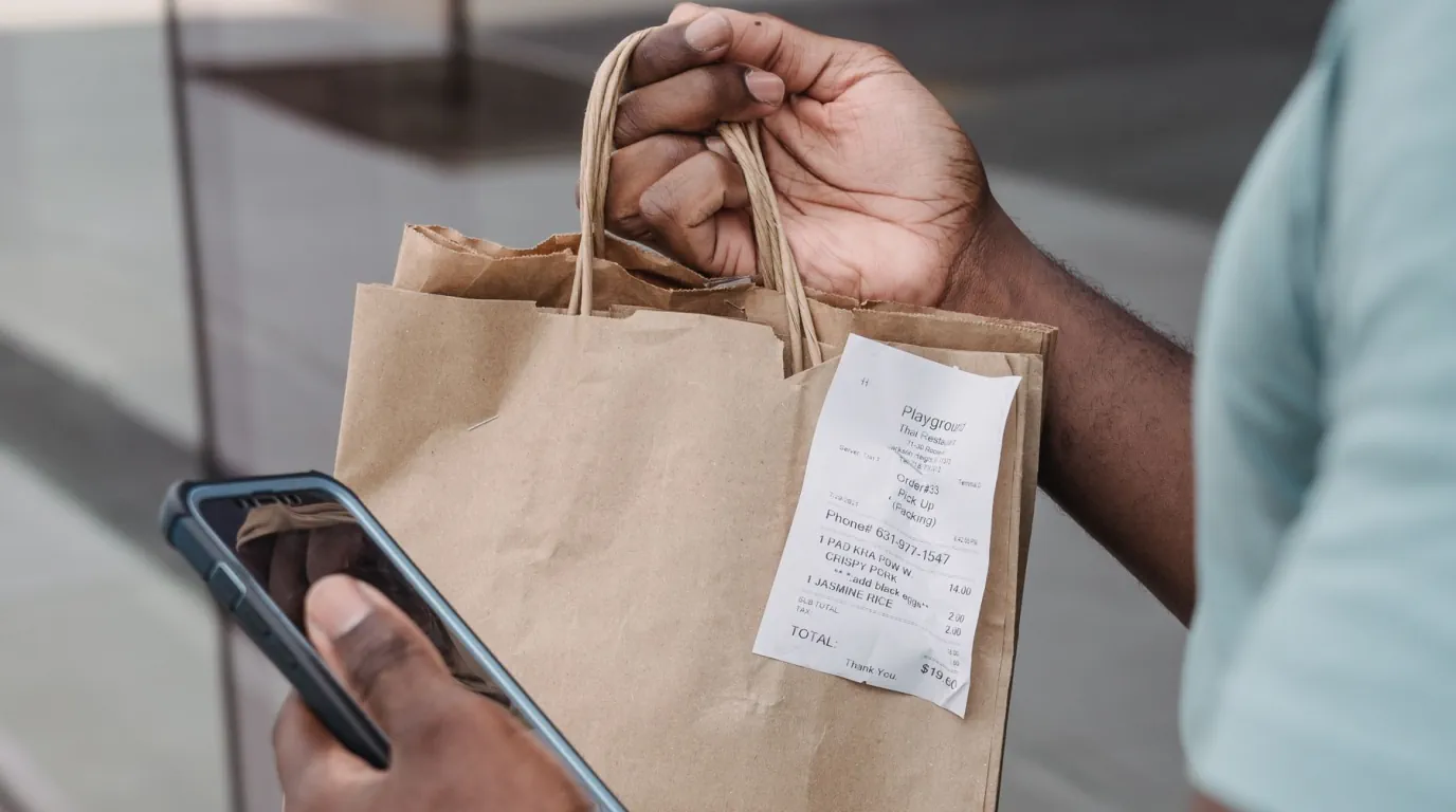A shopper checks their phone while holding a grocery bag and a long receipt.